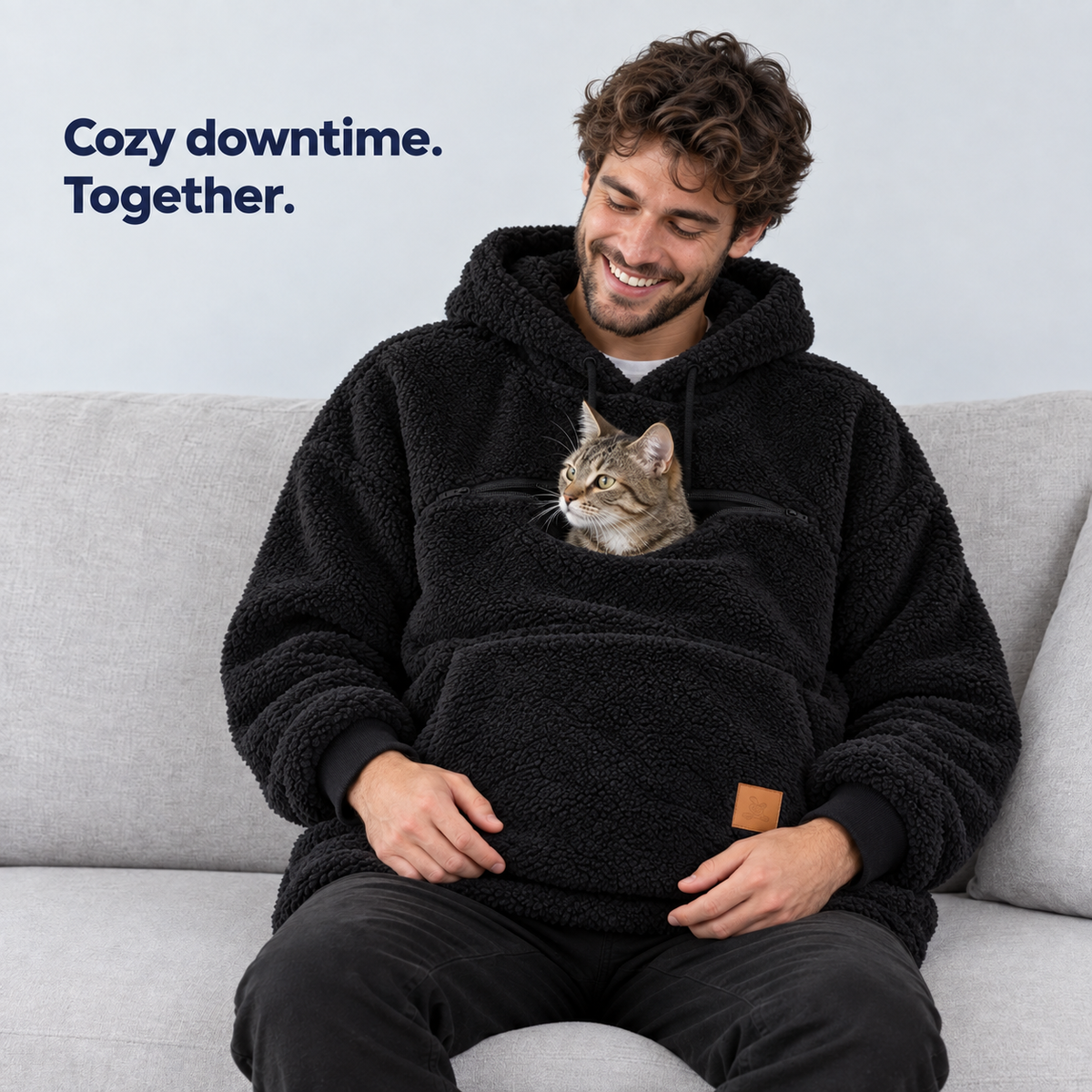 Man seated in the black sherpa pet carrier hoodie with a tabby cat in the pouch. Cozy downtime. Together.