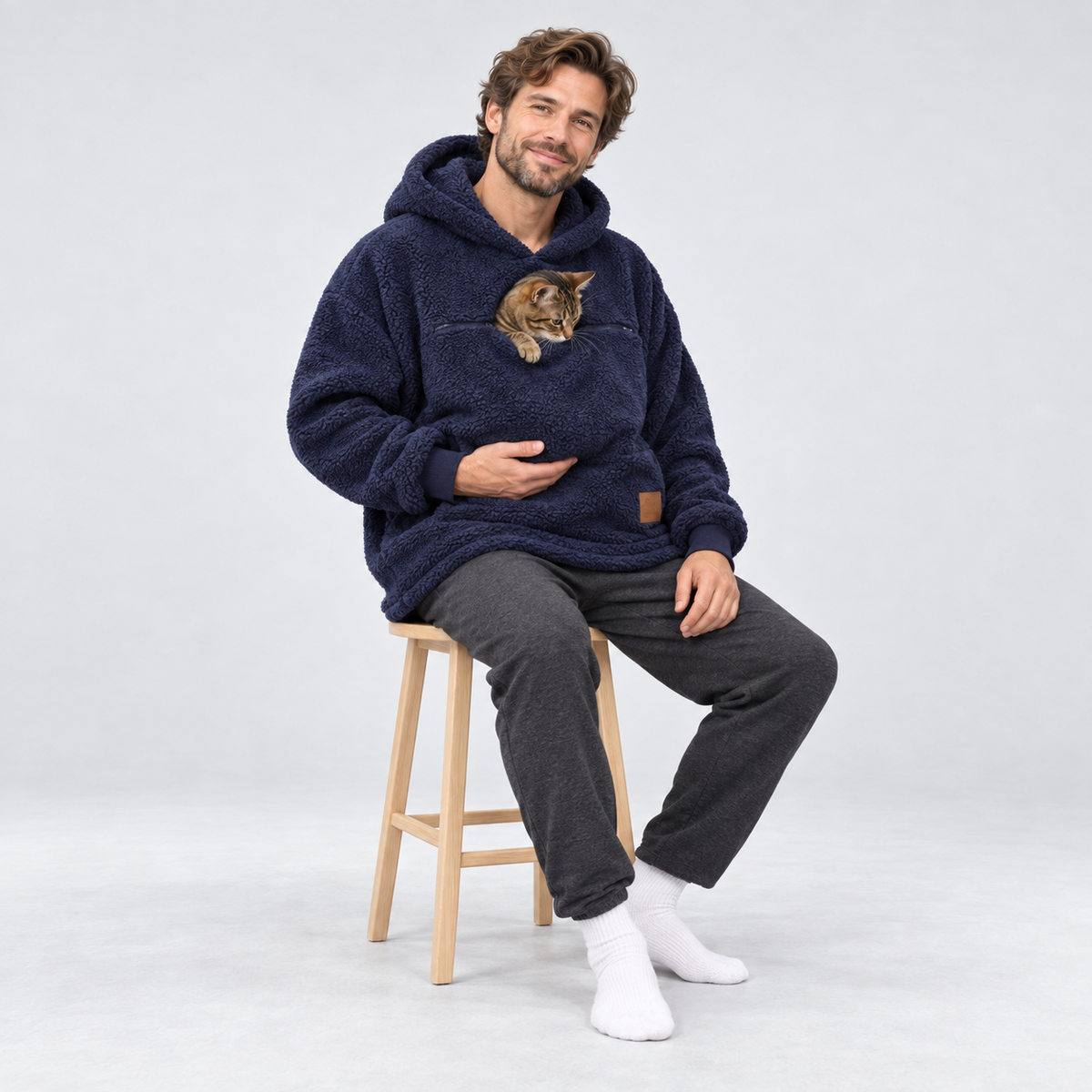 Navy color: man seated on a stool wearing the sherpa pet carrier hoodie with a tabby cat.