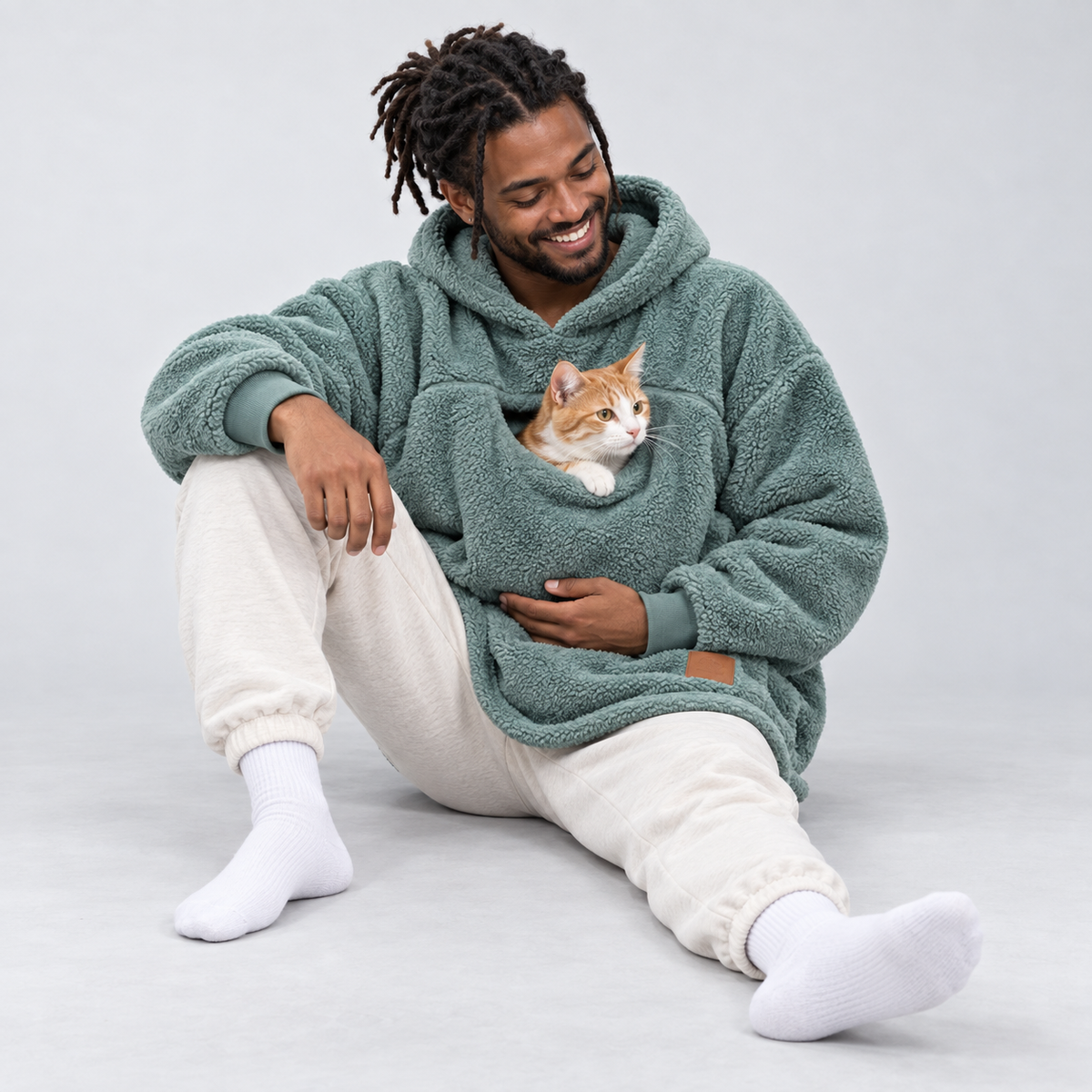 Cyan color: seated man wearing the sherpa pet carrier hoodie with an orange-and-white cat in the front pouch.